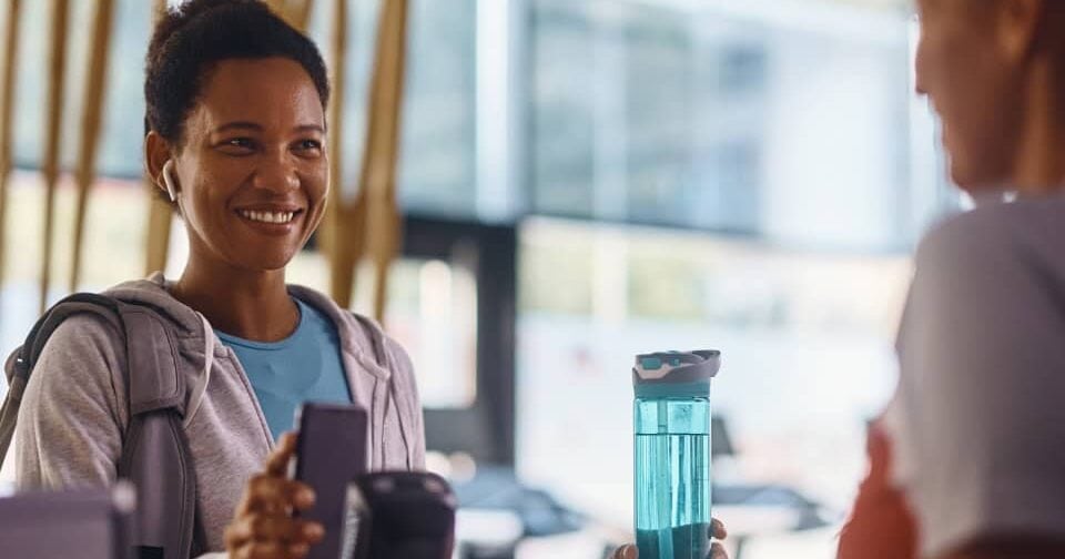 Una mujer sonriente en una recepción de gimnasio, sosteniendo una botella de agua azul y su teléfono móvil, mientras habla con otra persona al otro lado del mostrador. El ambiente es moderno y luminoso, con detalles del gimnasio en el fondo.