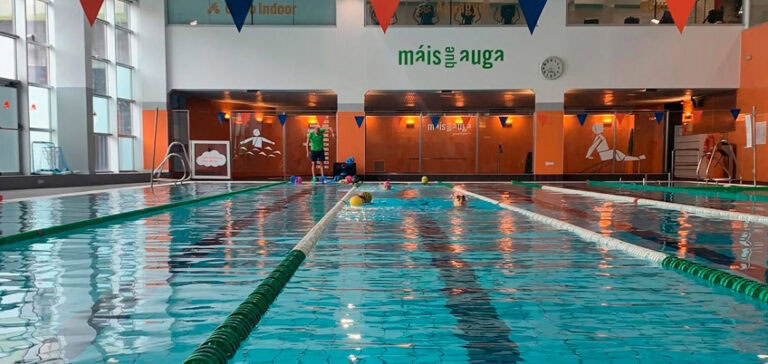 dividida en carriles con flotadores verdes, y se pueden ver algunos nadadores utilizando la piscina. A lo largo del borde de la piscina, hay varios objetos flotantes de colores y una persona de pie cerca de una puerta de vidrio. En el fondo, hay paredes anaranjadas decoradas con figuras de personas nadando y haciendo ejercicio, y un reloj en la pared superior derecha. El techo tiene banderines triangulares en colores azul y rojo, y las grandes ventanas permiten la entrada de luz natural, iluminando el área de la piscina.