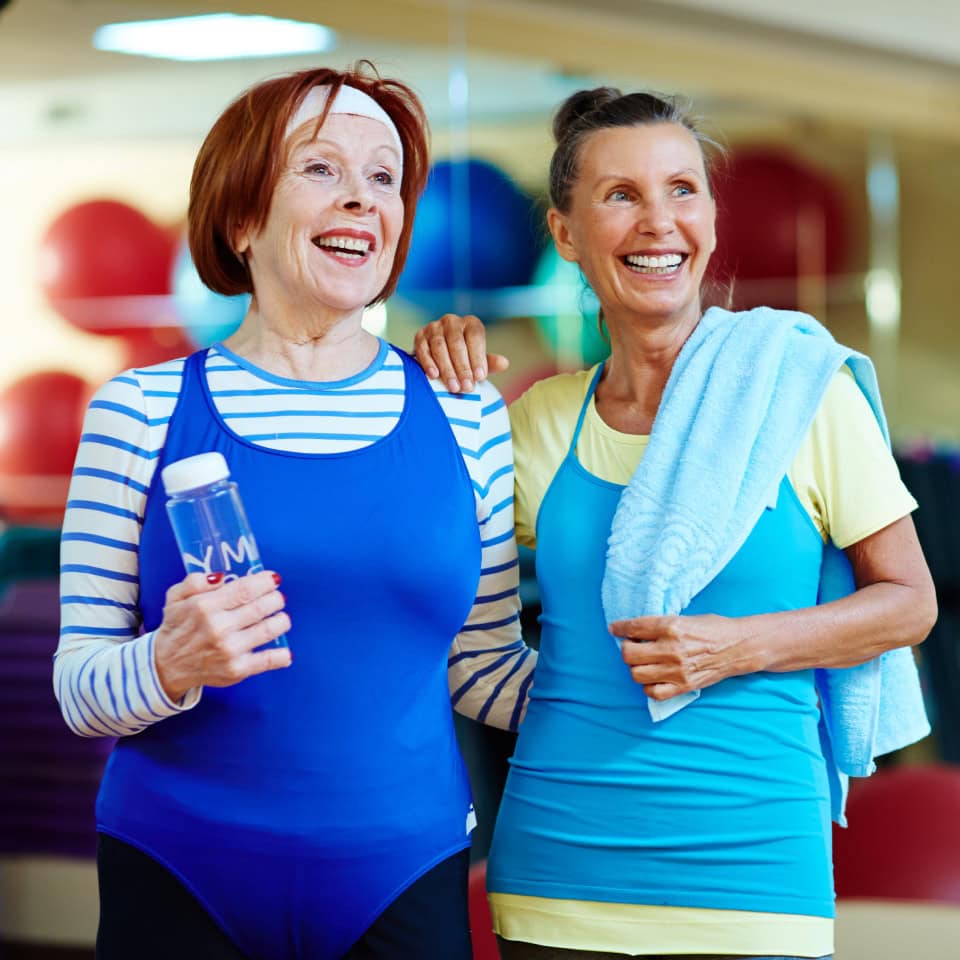 Dos mujeres mayores sonrientes en un gimnasio, vestidas con ropa deportiva azul. Una de ellas sostiene una botella de agua y la otra lleva una toalla sobre el hombro. Están de pie juntas en un entorno de gimnasio con balones de ejercicio de colores en el fondo.