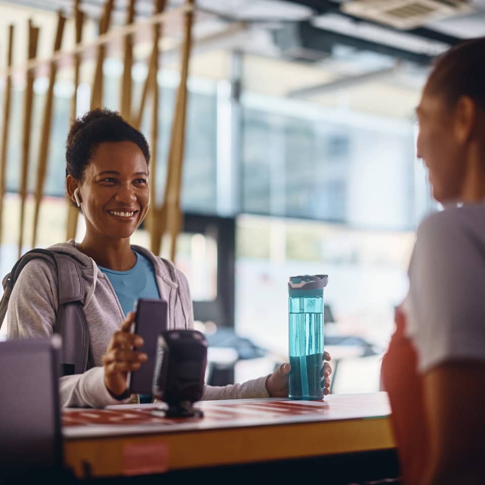 Una mujer sonriente en una recepción de gimnasio, sosteniendo una botella de agua azul y su teléfono móvil, mientras habla con otra persona al otro lado del mostrador. El ambiente es moderno y luminoso, con detalles del gimnasio en el fondo.