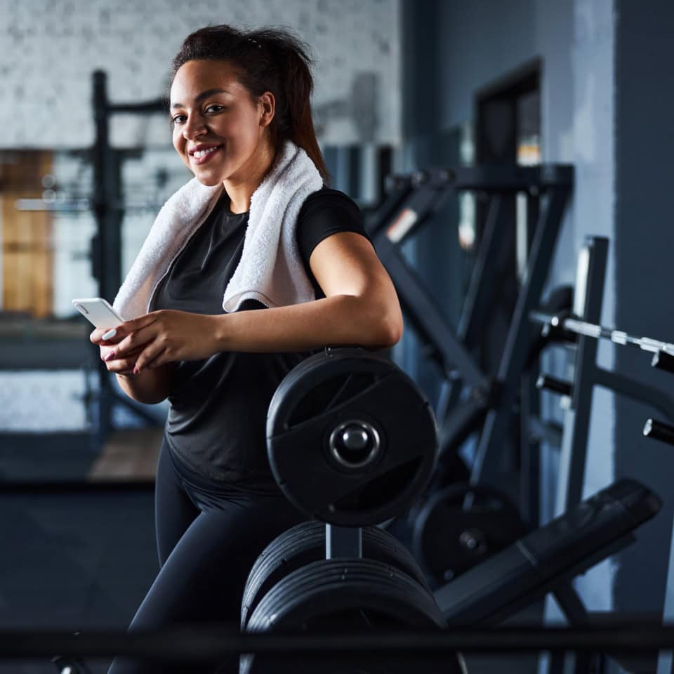 Mujer joven sonriente en un gimnasio, apoyada en una barra de pesas con una toalla blanca alrededor de su cuello y sosteniendo un teléfono móvil. El entorno es un área de entrenamiento con equipos de musculación visibles en el fondo.